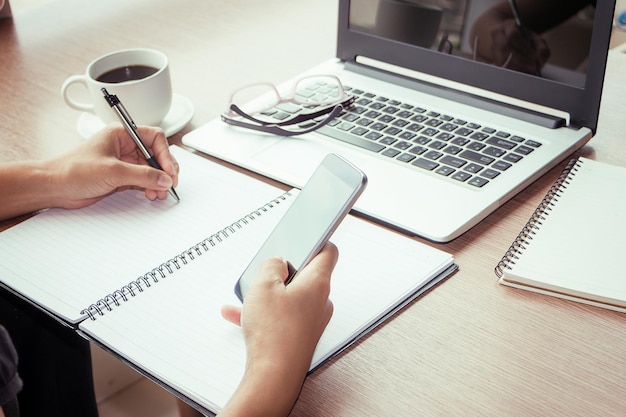 Image coupée de la main utilisant un téléphone portable tout en écrivant sur un livre en spirale avec un ordinateur portable et une tasse de café sur la table