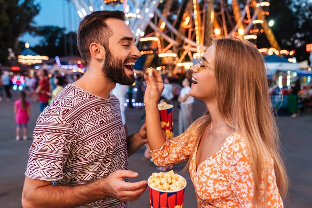 Image d'un couple d'amoureux souriant et positif marchant à l'extérieur dans un parc d'attractions mange du pop-corn.