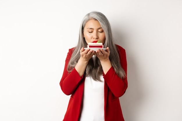 Image d'une dame senior asiatique drôle reniflant un délicieux gâteau, une odeur de dessert avec un visage satisfait, debout sur fond blanc.