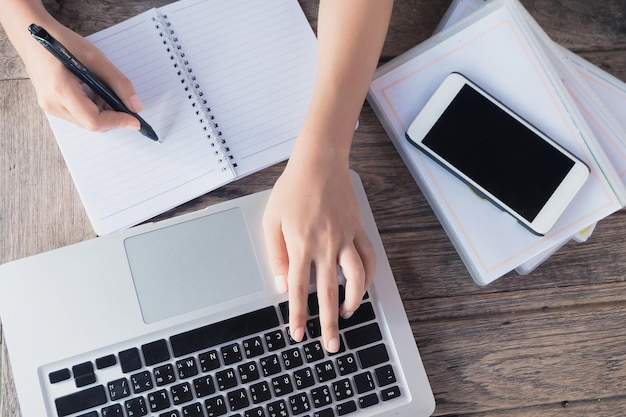 Image découpée d'une femme écrivant sur un livre tout en utilisant un ordinateur portable par téléphone à la table