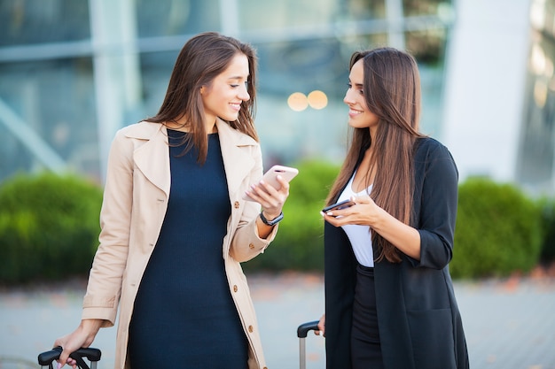 Image de deux européennes joyeuses regardant un smartphone, debout avec leurs bagages, près de l'aéroport, en attente du vol ou après le départ. Voyage en avion