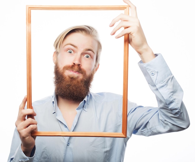 Image drôle. Jeune homme portant une barbe tenant un cadre photo devant son visage et un martèlement amusant, isolé sur blanc.