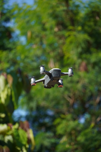 Image d'un drone blanc et gris volant, avec des arbres et du ciel autour.