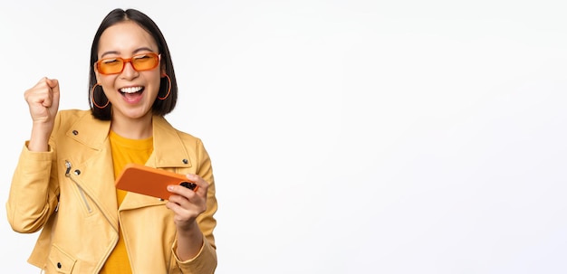 Image d'élégante fille coréenne dansant avec un smartphone riant heureux et souriant debout sur fond blanc