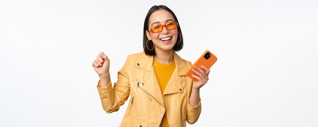 Image d'élégante fille coréenne dansant avec un smartphone riant heureux et souriant debout sur fond blanc