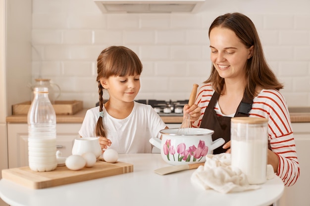 Image d'une enfant souriante aux cheveux noirs cuisinant avec sa mère une pâtisserie maison assise à table dans la cuisine exprimant des émotions positives et du bonheur