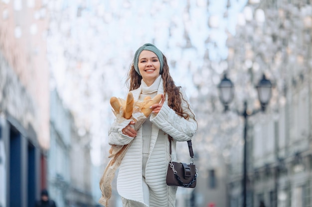 L'image est en mouvement. Jolie fille en costume blanc et bande bleue avec un sac sur son épaule tient plusieurs baguettes de pain. Arrière-plan flou du centre-ville. Image publicitaire avec place pour le logo.