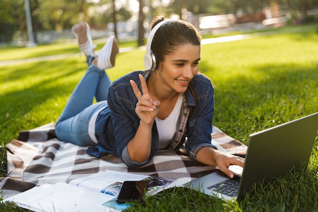 Image d'étonnante femme heureuse étudiante se trouve à l'extérieur dans un parc à l'aide d'un ordinateur portable, écouter de la musique faire un geste de paix.