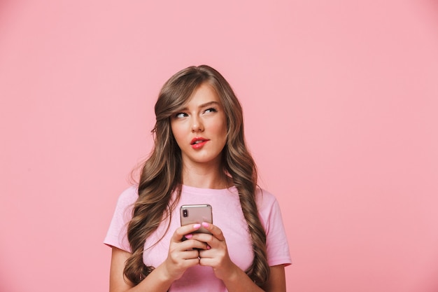 Image d'une femme adorable des années 20 avec de longs cheveux bouclés et un regard ludique tenant un téléphone mobile dans les mains tout en discutant, isolé sur fond rose