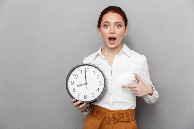 Image d'une femme d'affaires rousse émotionnelle des années 20 en tenue de soirée se demandant tout en tenant une grande horloge isolée sur fond gris