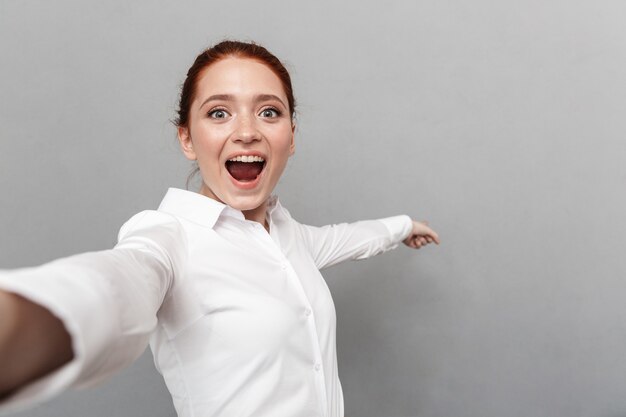 Image d'une femme d'affaires rousse heureuse de 20 ans en tenue de soirée souriante tout en prenant une photo de selfie isolée sur fond gris