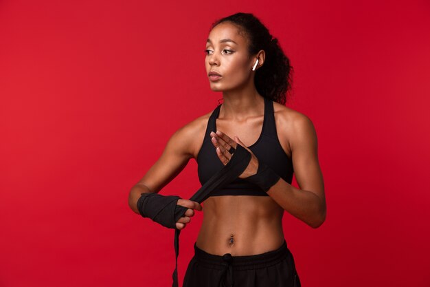 Image de femme afro-américaine sportive en vêtements de sport noir portant des bandages de sport sur ses mains, isolé sur mur rouge