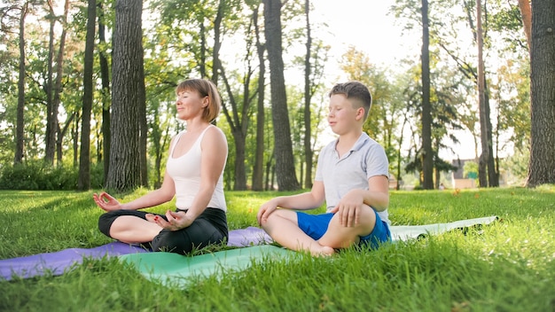 Image d'une femme d'âge moyen enseignant à un adolescent faisant du yoga et du fitness sur l'herbe au parc. La famille prend soin de sa santé