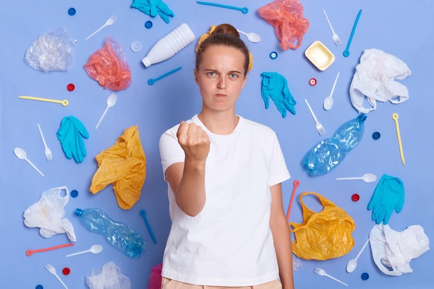 Image d'une femme agressive en colère vêtue d'un t-shirt blanc entourée de litière en plastique cueillie isolée sur un mur bleu montrant le poing à la caméra veut punir ceux qui nuisent à la nature