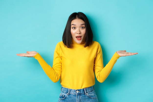 Image d'une femme asiatique indécise haussant les épaules, écartant les mains sur le côté et ayant l'air désemparée devant la caméra, debout confuse sur fond bleu