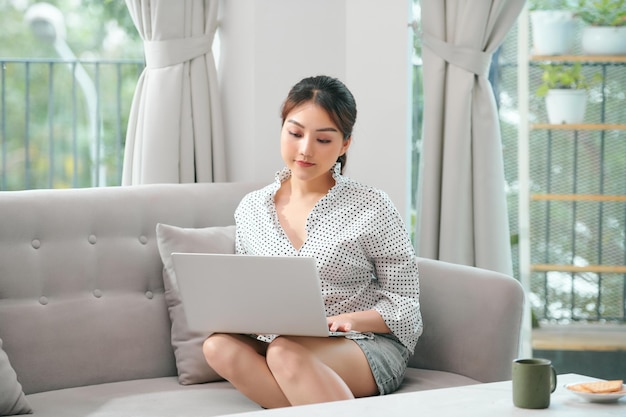 Image d'une femme asiatique joyeuse utilisant un ordinateur portable assise sur un canapé dans le salon