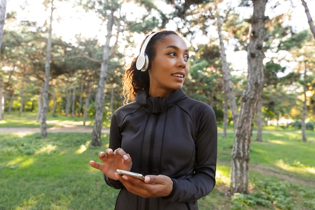 Image de la femme athlétique des années 20 portant un survêtement noir et des écouteurs, à l'aide d'un téléphone mobile tout en marchant dans le parc verdoyant