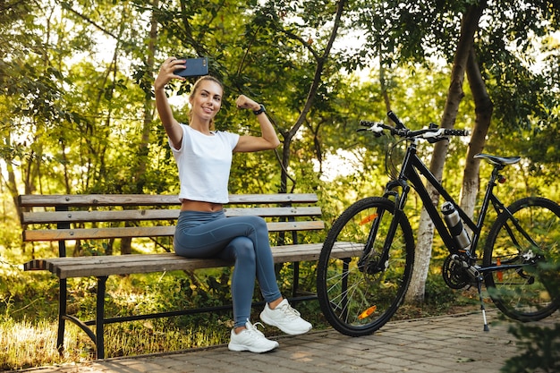 Image d'une femme athlétique assise sur un banc dans le parc à vélo et prendre une photo de selfie sur un téléphone portable