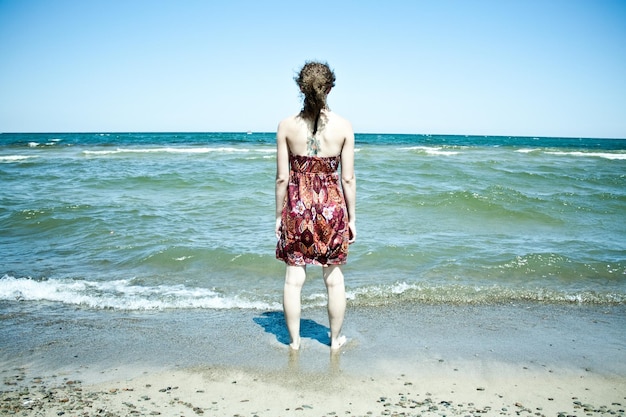 Image d'une femme aux longs cheveux bruns vêtue d'une robe d'été rouge se tient dans l'eau jusqu'aux chevilles sur la plage alors que les vagues clapotent sur ses chevilles