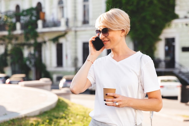 Image d'une femme blonde caucasienne portant un t-shirt blanc et des lunettes de soleil marchant dans la rue de la ville en été avec du café à emporter, et parlant au téléphone mobile
