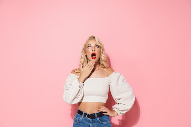 Image d'une femme blonde choquée aux longs cheveux bouclés exprimant l'émerveillement et regardant vers le haut le fond isolé sur un mur rose