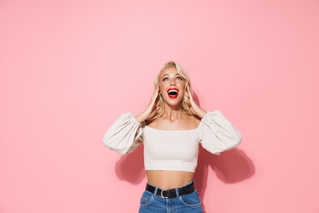 Image d'une femme blonde excitée aux longs cheveux bouclés portant des vêtements à la mode souriant et regardant vers le haut sur un fond isolé sur un mur rose