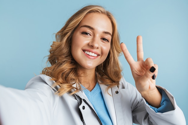 Image d'une femme blonde heureuse de 20 ans portant un imperméable souriant tout en prenant une photo de selfie