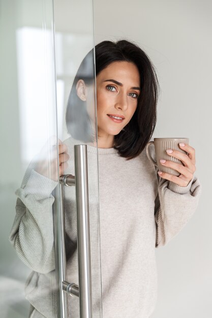 Image de femme brune 30 s tenant une tasse de thé, en se tenant debout dans une salle lumineuse moderne le matin