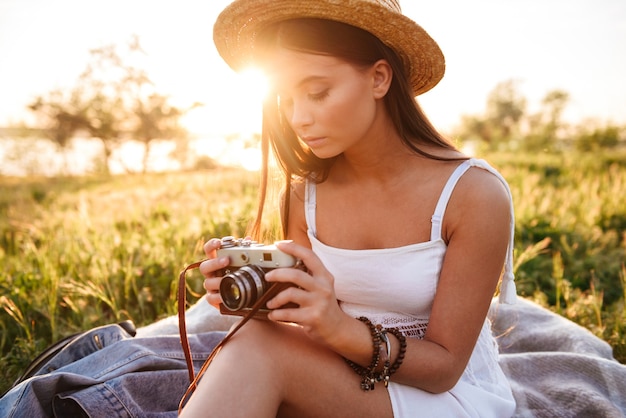 Image de femme brune européenne aux cheveux longs portant un chapeau de paille et une robe blanche tenant un appareil photo rétro alors qu'il était assis sur l'herbe dans le parc pendant les loisirs