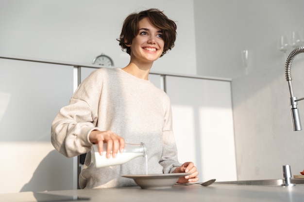 Image d'une femme brune heureuse en train de dîner en se tenant debout près de la table à la cuisine