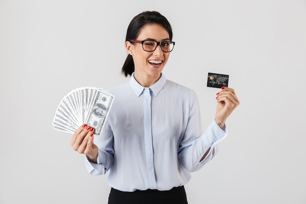 Image de femme de bureau réussie portant des lunettes tenant un tas d'argent et de carte de crédit, isolé sur un mur blanc
