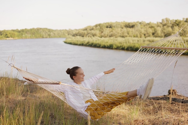 Image D'une Femme Caucasienne Souriante Et Ravie Portant Une Chemise Blanche Assise Sur Un Hamac Au Bord De La Rivière Regardant La Belle Nature En Appréciant Le Repos