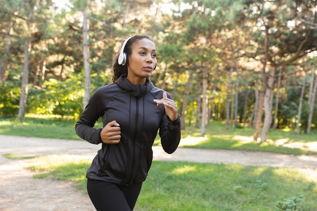 Image d'une femme énergique de 20 ans portant un survêtement noir et des écouteurs, tout en courant dans le parc verdoyant