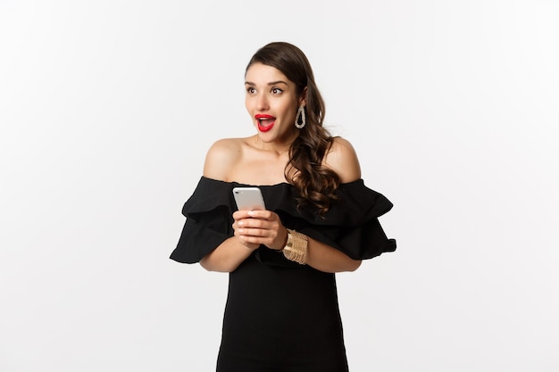 Image d'une femme excitée en robe glamour noire, rouge à lèvres rouge, utilisant un téléphone portable et regardant à gauche étonnée, debout sur fond blanc.