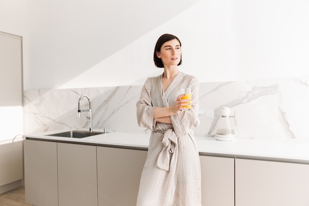 Image de femme fascinante aux cheveux noirs courts debout dans la cuisine et boire du jus d'orange, à partir de verre transparent