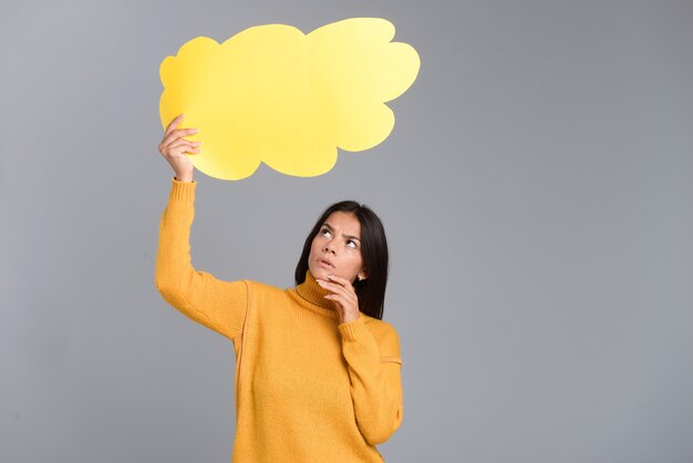 Image d'une femme pensante posant isolée sur un mur gris tenant une bulle de pensée.