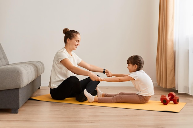 Image d'une femme avec une petite fille portant des vêtements de sport faisant des exercices de sport à la maison assise sur le sol tenant les mains ensemble et pratiquant le yoga à la maison faisant des exercices d'étirement