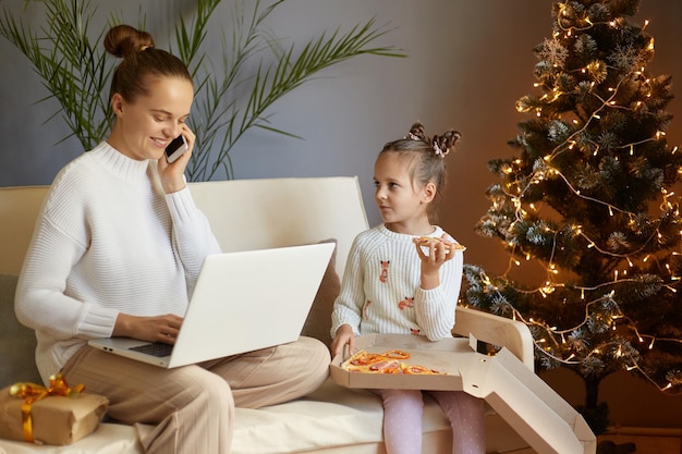 Image d'une femme de race blanche avec une coiffure en chignon assise travaillant sur un ordinateur portable et parlant au téléphone portable sa jolie fille charmante mangeant de la pizza et jouant près de sa mère