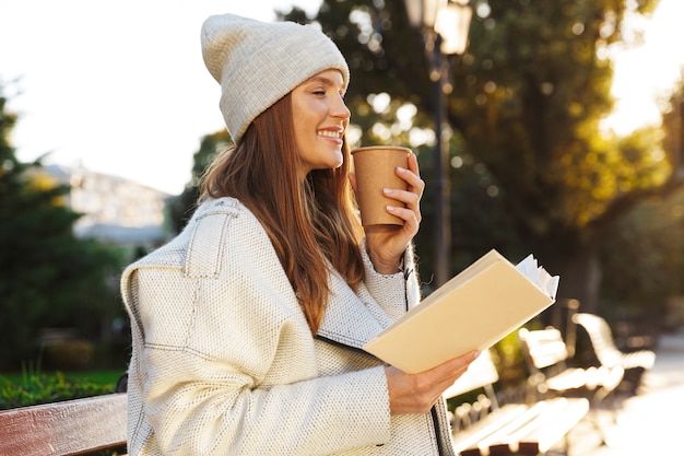 Image D'une Femme Rousse Assise Sur Un Banc à L'extérieur Tenant Un Livre De Lecture, Boire Du Café.