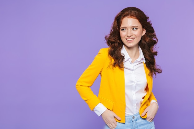 Image de femme rousse européenne portant une veste jaune souriant et regardant de côté à copyspace over purple