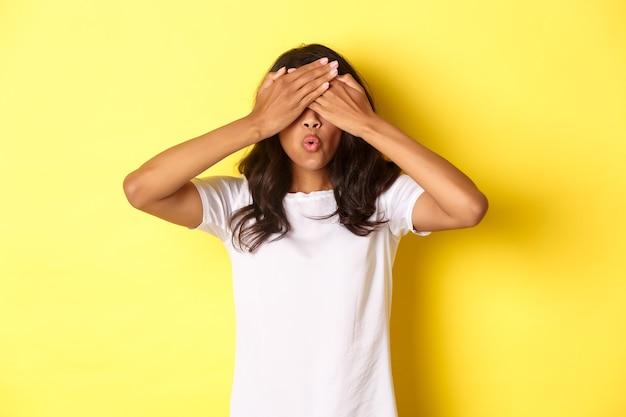 Image d'une fille afro-américaine excitée attendant la surprise, souriante et couvrant les yeux avec les mains, debout sur fond jaune.