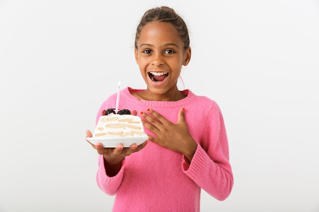 Image d'une fille afro-américaine ravie tenant un morceau de tourte avec une bougie et souriante isolée sur un mur blanc
