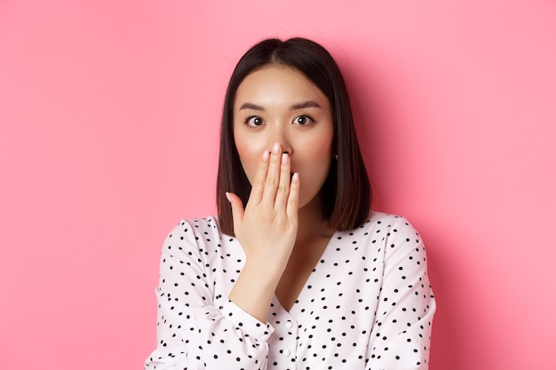 Image d'une fille asiatique choquée bavardant, haletant et couvrant la bouche, regardant la caméra avec une incrédulité totale, debout sur fond rose