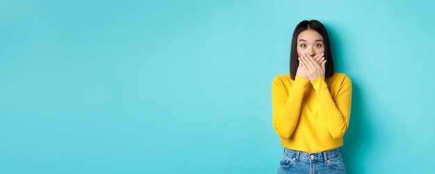 Image d'une fille asiatique choquée se couvrir la bouche et regarder la caméra portant un chandail jaune contre une ba bleue