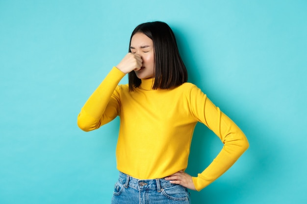 Image d'une fille asiatique élégante fermant le nez et grimaçant d'aversion, quelque chose sentait mauvais, debout dégoûté sur fond bleu.