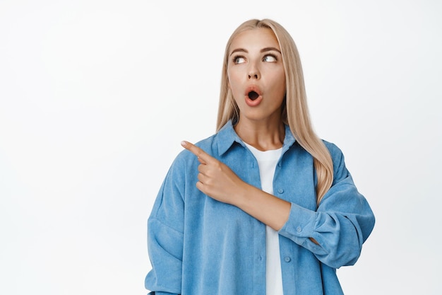 Image d'une fille blonde surprise disant wow pointant et regardant le coin supérieur gauche avec une expression de visage impressionné debout dans des vêtements décontractés sur fond blanc