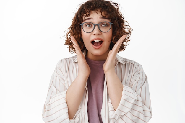 Image D'une Fille Bouclée Surprise Haleter Et Dire Wow, Regarder Avec Enthousiasme Une Publicité Incroyable Et Cool, Vérifier L'offre Promotionnelle, Debout Dans Des Verres Et Des Vêtements Sur Fond Blanc