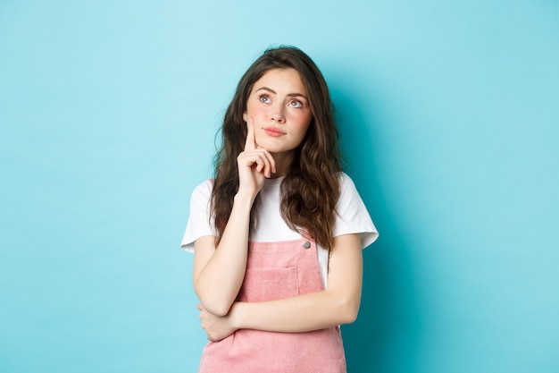 Image d'une fille glamour mignonne et réfléchie avec du maquillage, regardant le coin supérieur gauche pensif, pensant ou imaginant quelque chose, debout sur fond bleu.