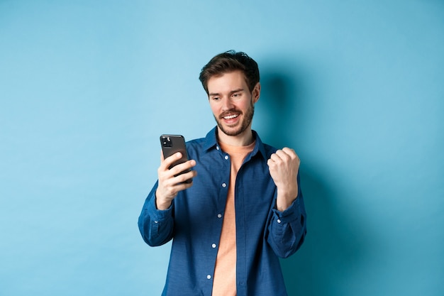 Image d'un gars chanceux gagnant en ligne, lisant l'écran du téléphone mobile et faisant la pompe de poing, triomphant, debout sur fond bleu.