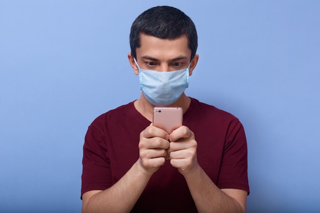 Image d'un gars curieux choqué tenant un téléphone mobile dans les deux mains, vérifiant les nouvelles, recherchant du contenu, paniqué, lisant des informations sur covid19, portant un masque antibactérien. Concept de coronavirus.